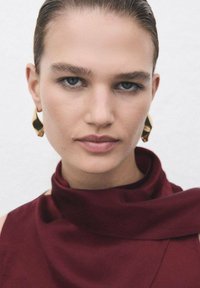 Prenda burdeos de cuello alto con textura suave, que presenta un diseño drapeado. La modelo tiene el cabello liso y lleva pendientes de aro dorados.