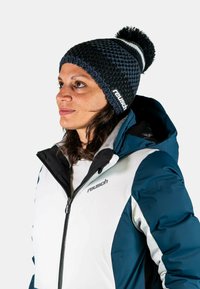 Knitted navy beanie with a pom-pom and white stripe, worn with a white and teal insulated jacket featuring a hood and zip closure.
