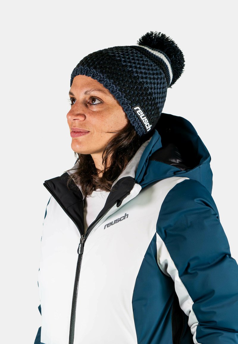 Knitted navy beanie with a pom-pom and white stripe, worn with a white and teal insulated jacket featuring a hood and zip closure.