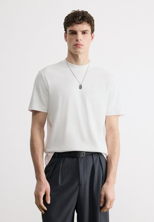 Young man with short curly hair wearing a plain white t-shirt, black belt, dark tailored pants, and a silver pendant necklace.