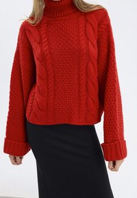 Pull en tricot rouge à col haut, avec des motifs en torsades et des manches texturées. Porté avec une jupe noire.