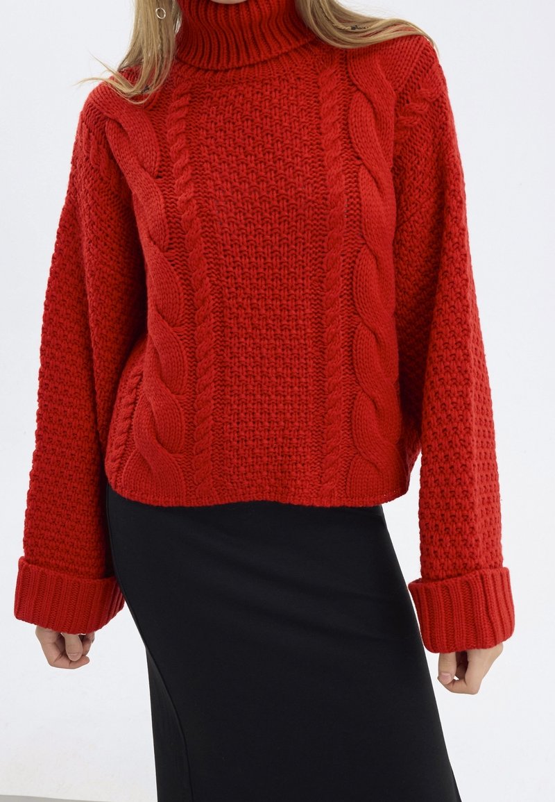 Pull en tricot rouge à col haut, avec des motifs en torsades et des manches texturées. Porté avec une jupe noire.
