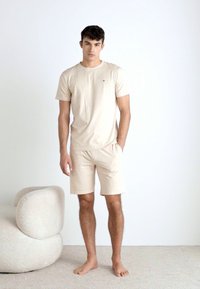 Camiseta de algodón beige de manga corta, combinada con pantalones cortos beige a juego. Diseño sencillo, corte relajado, textura suave, de pie sobre un fondo neutro.