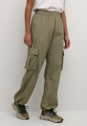 Personne portant un pantalon cargo vert olive avec des chevilles élastiques et des poches latérales, assorti à des baskets beiges et vertes et une chemise blanche.