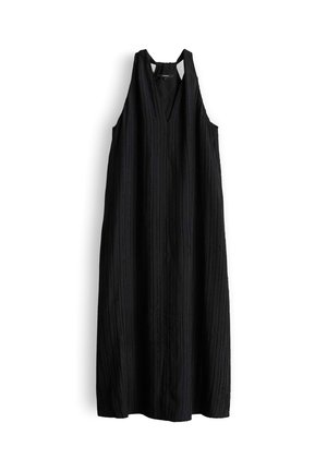 Frau steht mit den Händen hinter dem Rücken, trägt ein ärmelloses schwarzes plissiertes Midikleid und schwarze Sandaletten mit Absatz vor weißem Hintergrund.