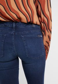 Jeans de mezclilla azul oscuro con un diseño ajustado y una textura sutil. Los bolsillos traseros están cosidos, y la cinturilla presenta una etiqueta y un detalle de botón.