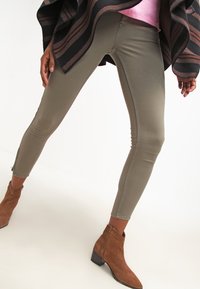 Dopasowane oliwkowe dżinsy z subtelnymi detalami w stębnowaniu i bocznymi zamkami, zestawione z brązowymi botkami za kostkę i przewiązane ciemnym w długi pas.