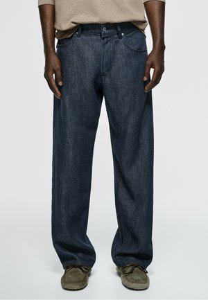 Uomo che indossa jeans blu scuro larghi, scarpe stringate in camoscio marrone e camicia beige a maniche lunghe, in piedi contro uno sfondo neutro.