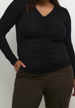 Femme portant un haut noir froncé à manches longues avec un col en V et un pantalon vert olive, debout avec une main dans la poche.