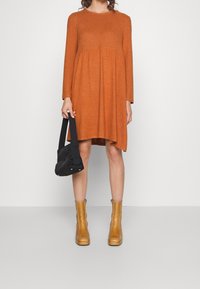 Vestido naranja quemado de manga larga con dobladillo acampanado y cintura fruncida. Combinado con un bolso negro y botines tobilleros color trigo. Presenta una textura suave.
