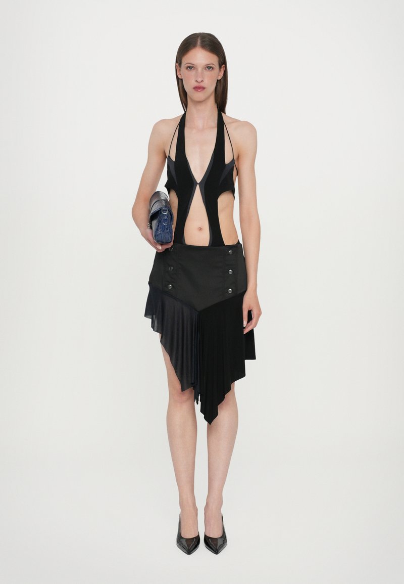 Black halter neck bodysuit with cut-out sides paired with a high-low black skirt featuring pleats and button details. Model holds a clutch.