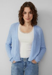 Lichtblauwe gebreide cardigan met open voorkant, geribbelde textuur, lange mouwen en een ontspannen pasvorm, gedragen over een witte tanktop.