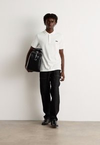 Weißes Poloshirt mit Kragen, kombiniert mit dunkelblauen Denim-Hosen. Das Model trägt eine schwarze Ledertasche und steht auf einem hellen Boden.