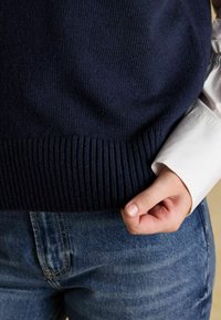 Jūras spēku adīta veste ar ribotu apakšdaļu, apvienota ar baltu kreklu. Redzama roka, kas tur kreklu pie aproces, stilizēta ar zilo džinsu biksēm.