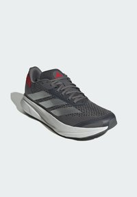 Zapatilla deportiva gris con parte superior de malla, acentos rojos y tres franjas blancas. Cuenta con una suela acolchada y un área de cordones texturizada para mayor soporte.