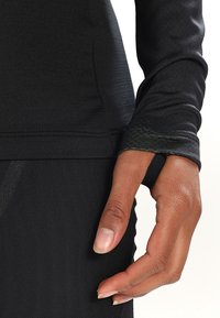 Haut de sport noir à manches longues avec tissu extensible. Comprend un trou pour le pouce au niveau du poignet, une texture lisse et une coupe ajustée.