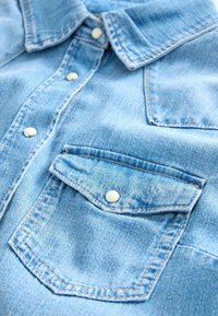 Gros plan sur une chemise en denim bleu clair montrant des poches avant boutonnées et des boutons-pression blancs sur la patte de boutonnage et le col.