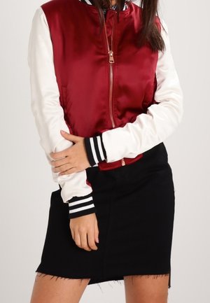 Personne portant une veste universitaire rouge et blanche avec des poignets rayés noirs et blancs et une jupe noire à ourlet brut.