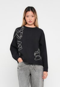 Ung kvinne med langt hår som har på seg svart hettegenser med hvit abstrakt linjekunst og lys gråkledde jeans, stående mot en ensfarget hvit bakgrunn.