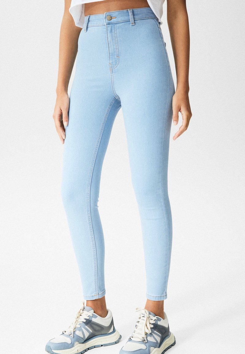 Jeans ajustados de talle alto en azul claro, hechos de denim, con detalles cosidos y un cierre clásico de botón. Combinados con zapatillas grises y blancas.
