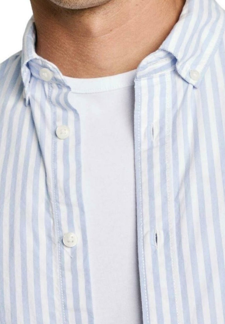 Primo piano di una persona che indossa una maglietta bianca a girocollo sotto una camicia a bottoni azzurro chiaro e bianca con i bottoni aperti.