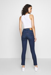 Jeans de tiro alto de color azul oscuro con un diseño ajustado, cinco bolsillos y un detalle de parche de cuero en la cinturilla. Top corto blanco.