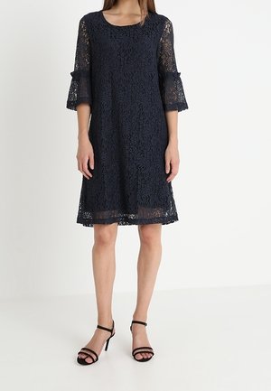 Nainen, jolla on polvipituinen tummansininen pitsimekko, jossa on kolmen neljäsosan pituinen kellohihat, sekä mustat remmisandaalit korkeilla koroilla.