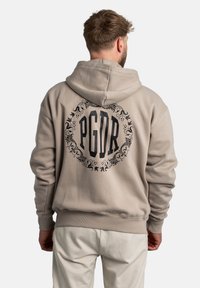 Sudadera beige con un gran gráfico negro de PGDR rodeado de un detallado ornamental en la parte posterior. Presenta una capucha con cordón y puños acanalados.