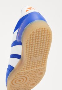 Zapatilla azul y blanca en ángulo para mostrar la suela de goma texturizada y el talón con logo naranja y el texto "EST. 1994".