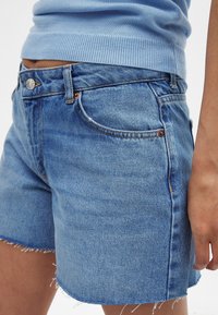 Šviesiai mėlynos džinsinės šortai su nusidėvėjusiu kraštu, turintys klasikinį penkių kišenių dizainą ir metalinį sagčių užsegimą. Audinio tekstūra atrodo minkšta ir nusidėvėjusi.