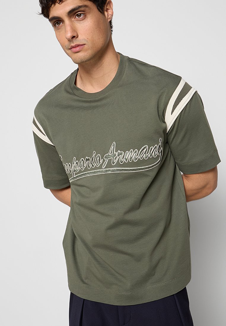 Joven con una camiseta Emporio Armani verde oliva con rayas blancas en los hombros, de pie con las manos detrás de la espalda sobre un fondo liso.