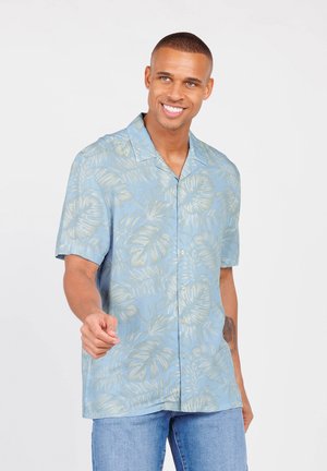 Camisa de manga corta de color azul claro con un patrón de hojas tropicales en color crema, diseño de botones y corte relajado, combinada con jeans de mezclilla azules.