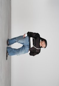 Γυναίκα που φοράει φαρδιά μπλε τζιν, μαύρο πουλόβερ, μαύρες σαγιονάρες και μία λευκή τσάντα χιαστί, μπροστά σε απλή γκρι απεικόνιση.