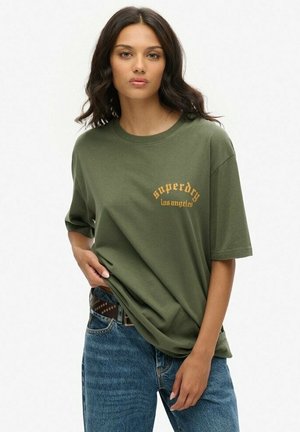 Oliwkowa bawełniana koszulka z luźnym krojem, okrągłym dekoltem i złotym nadrukiem "superdry los angeles" z przodu. Noszona z niebieskimi dżinsami.