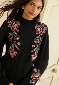 Fekete pulóver magas nyakkal, színes virágmintázattal rózsaszín, zöld és krémszínben az ujjakon és az elülső részen. Puha tapintású.