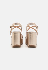 Tacchi a piattaforma metallici oro con cinturini incrociati alla caviglia e un tacco quadrato spesso. Texture liscia e design minimalista, incentrato su linee eleganti.