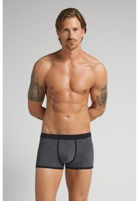 Muž bez košile s hnědými vlasy a tetováním na obou pažích, oblečený v tmavě šedých boxerkách s černým lemováním, stojící před šedým pozadím.