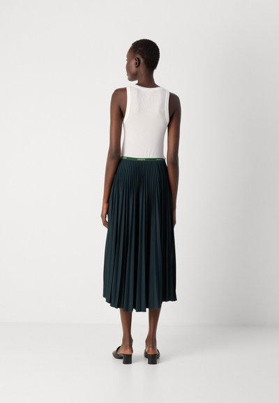 Top blanc sans manches associé à une jupe plissée vert foncé avec une ceinture accentuée de vert. Le modèle porte des chaussures à enfiler noires.