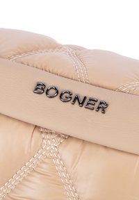 Beige tikattu laukku, jossa on sileä nahkainen hihna, johon on kohopainettu musta "BOGNER"-logo. Tarkka ompelu luo kohokuvioidun kuosin.