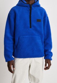 Pullover in pile blu con cappuccio, mezza zip e tasca frontale. Presenta una superficie morbida e texture e una piccola etichetta nera sul petto.