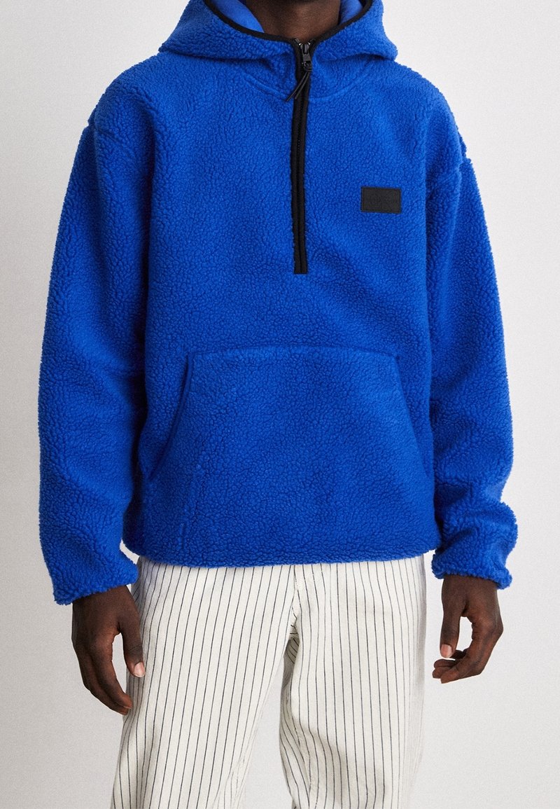 Pullover in pile blu con cappuccio, mezza zip e tasca frontale. Presenta una superficie morbida e texture e una piccola etichetta nera sul petto.