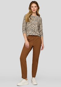 Langärmliges, gemustertes Oberteil in Creme und Tan mit rundem Halsausschnitt, kombiniert mit schmal geschnittenen braunen Hosen mit seitlichen Reißverschlüssen und einem Kordelzug.