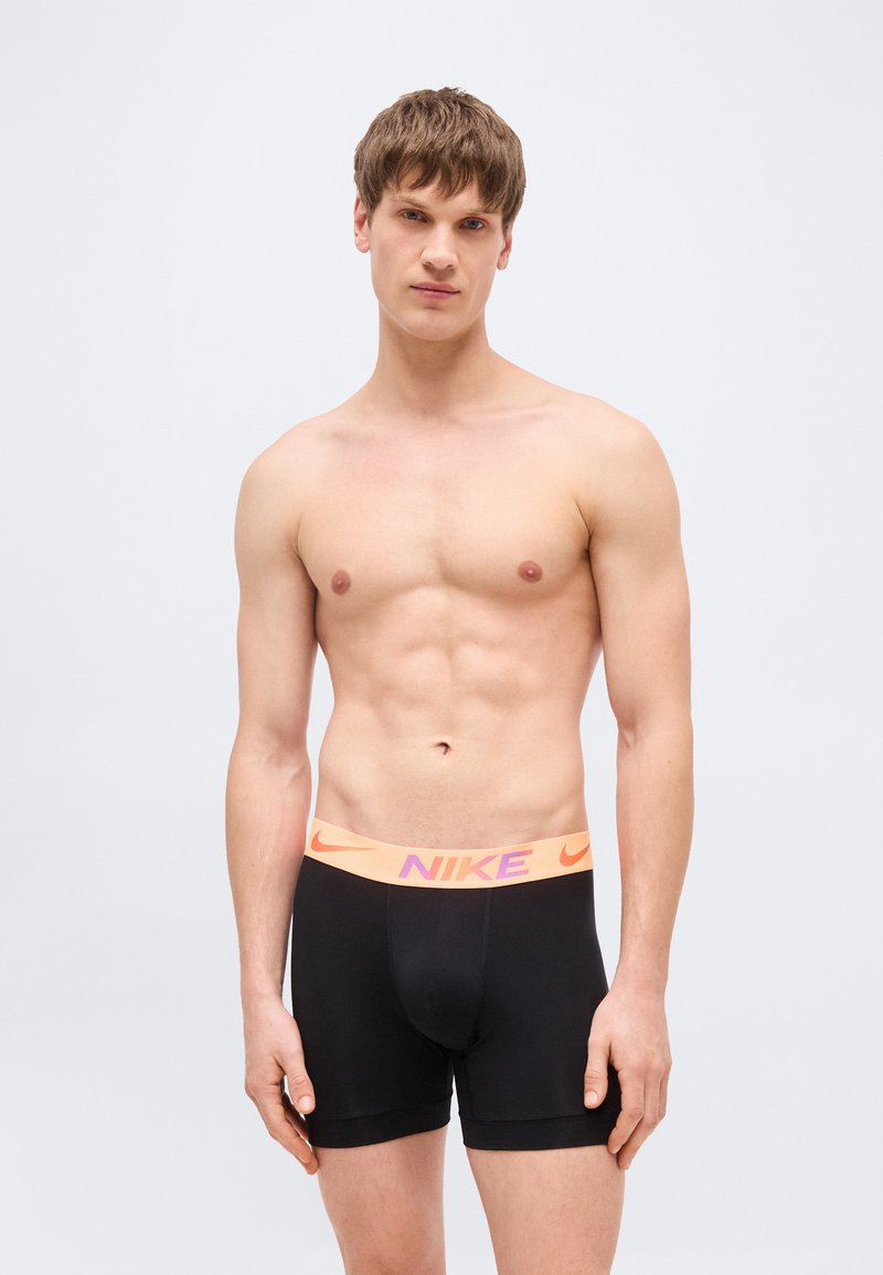 Jonge man met kort bruin haar, gekleed in zwarte Nike boxershorts met een oranje tailleband, op een effen lichtgrijze achtergrond.