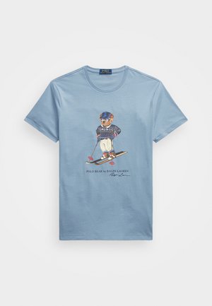 Vaaleansininen pyöreäkauluksinen t-paita, jossa karhu pukeutuneena hiihtovarusteisiin ja hiihtosauvat kädessään, alla teksti "Polo Bear by Ralph Lauren".