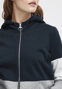Haut à capuche zippé bleu marine avec un design en blocs de couleur gris et blanc, doté d'une capuche à cordon de serrage et de détails en fermeture éclair argentés. Texture de tissu douce.