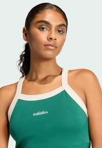 Zelený tielko s béžovými ramienkami a vyšívaným logom "adidas". Tkanina vyzerá hladko; výstrih je zaoblený. Jednoduchý dizajn.