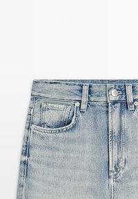 Jean en denim bleu clair, zone de la taille montrant les poches avant, passants de ceinture, bouton et braguette sur fond blanc.