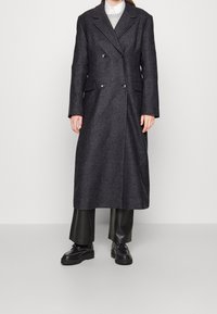 Abrigo de mezcla de lana gris oscuro, de doble botonadura, con solapas de muesca, bolsillos delanteros y una silueta larga y recta, que se lleva sobre pantalones y zapatos negros.