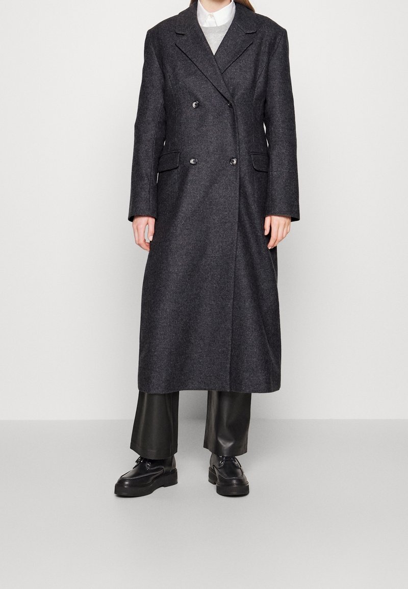 Abrigo de mezcla de lana gris oscuro, de doble botonadura, con solapas de muesca, bolsillos delanteros y una silueta larga y recta, que se lleva sobre pantalones y zapatos negros.