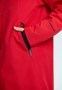 Giacca impermeabile rossa con una texture liscia, dotata di una tasca laterale con cerniera nera e un logo sulla manica.